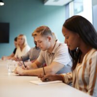 Group Of Young Candidates Sitting At Boardroom Table Making Notes At Presentation At Business Graduate Recruitment Assessment Day