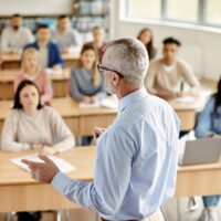 Back view of mature professor giving lecture to large group of college students in the classroom.